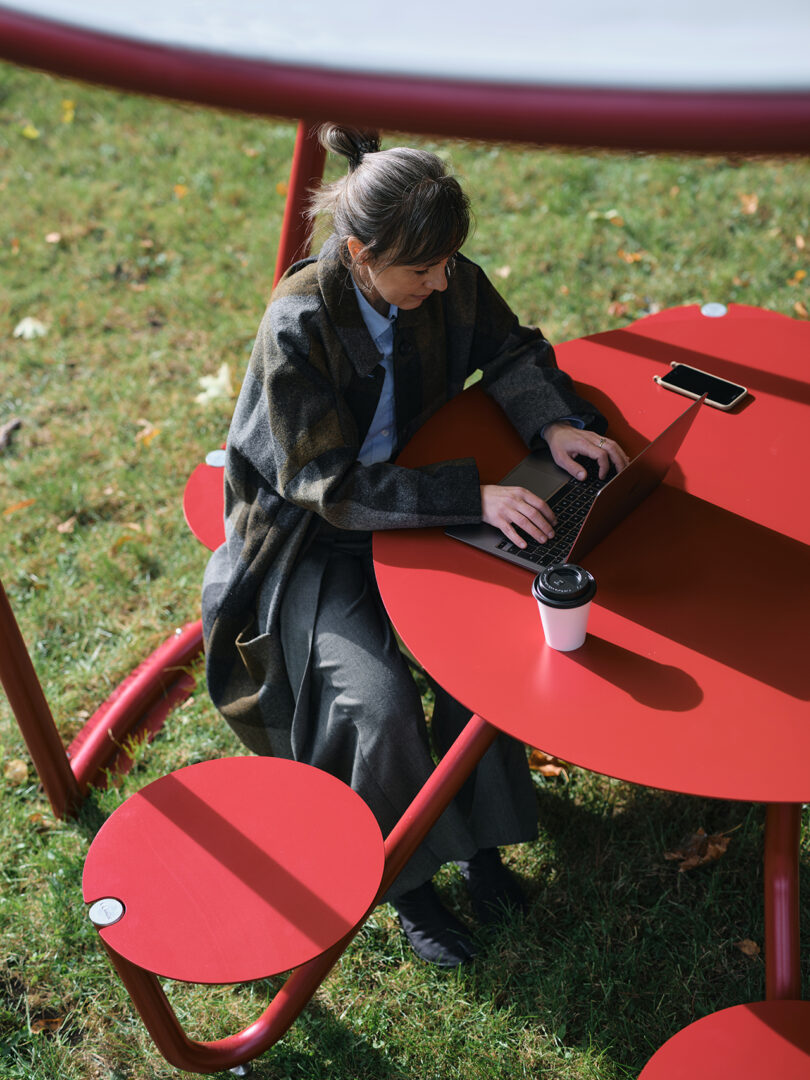 A woman sits at a red outdoor table in a park with lush green trees in the background.