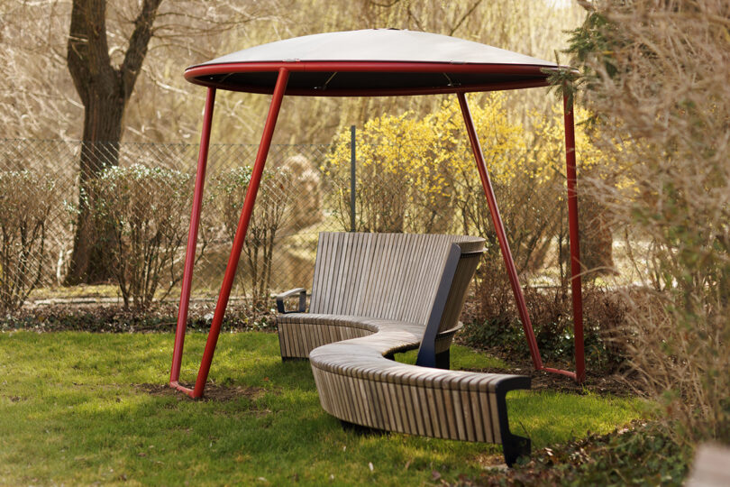 A curved wooden bench with a red canopy, surrounded by lush greenery and yellow blooming bushes.