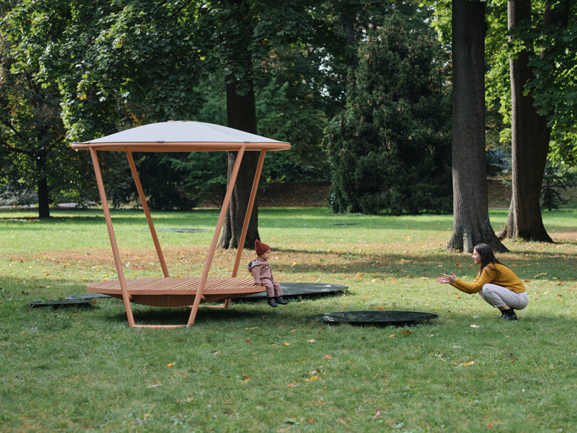 A woman and child playing on a modern wooden circular bench with an overhead canopy in a park setting, surrounded by green grass and trees.