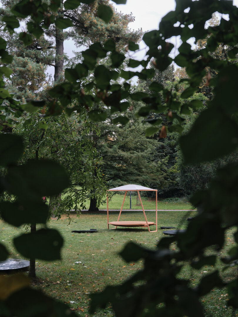 Modern wooden circular bench with an overhead canopy in a park setting, surrounded by green grass and trees.