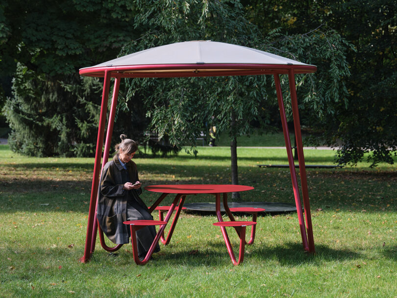 A woman sits at a red outdoor table in a park with lush green trees in the background.